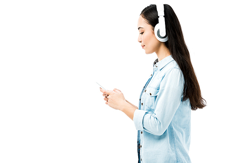 side view of attractive asian woman in denim shirt listening music and holding smartphone isolated on white