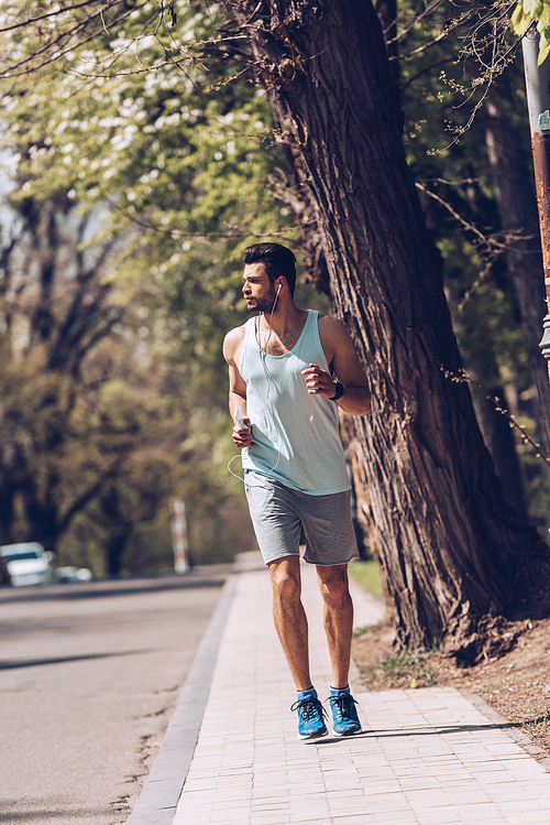 handsome sportsman listening music in earphones while jogging alone pavement near roadway