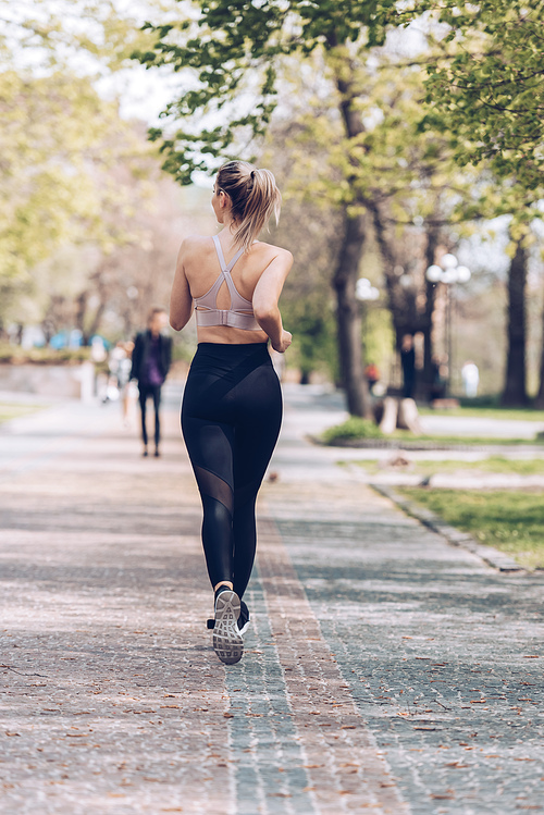 back view of young sportswoman jogging along wide walkway in park
