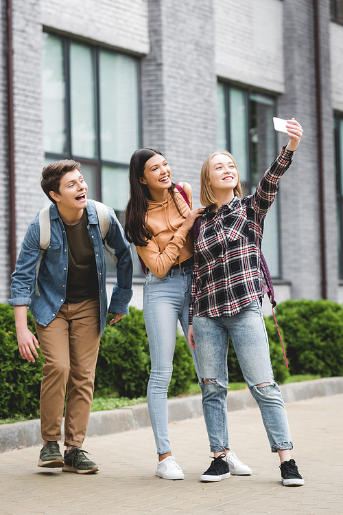 happy teenagers holding smartphone, taking selfie and smiling outside