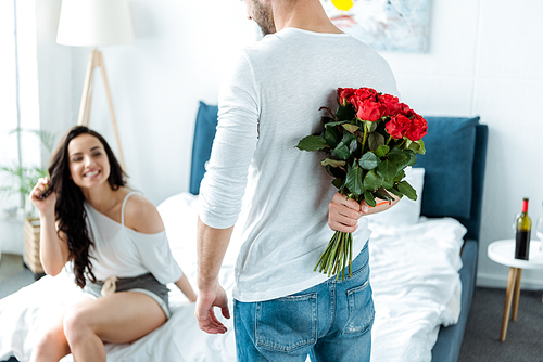 selective focus of happy girlfriend looking at boyfriend with red roses behind back