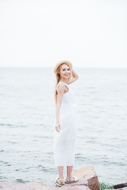 smiling blonde young woman touching straw hat and  while standing in white dress near sea