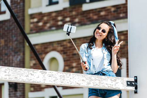 attractive woman holding selfie stick and taking selfie on smartphone while showing peace sign