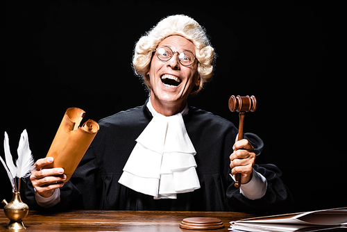 smiling judge in judicial robe and wig sitting at table and holding document and gavel isolated on black