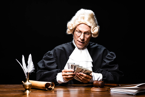 judge in judicial robe and wig sitting at table and counting money isolated on black