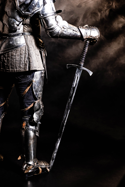 cropped view of knight in armor holding sword on black background