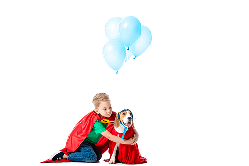 preschooler child  in red hero cloak embracing beagle dog near blue party balloons isolated on white