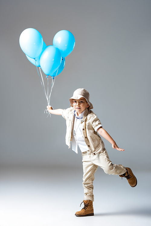 full length view of explorer child in glasses standing on one leg andholding blue balloons on grey