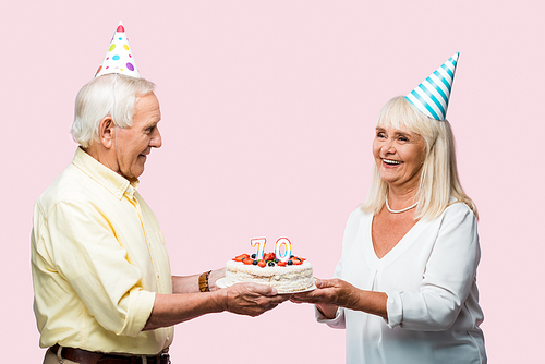 positive senior couple with grey hair holding birthday cake isolated on pink