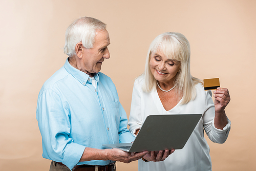 cheerful retired woman holding credit card near senior husband with laptop isolated on beige