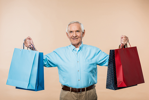 cheerful senior man smiling while holding shopping bags isolated on beige