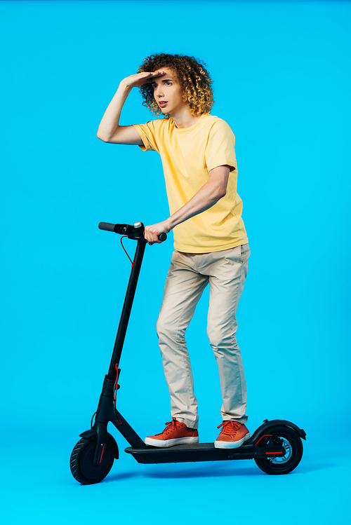 full length view of curly teenager riding electric scooter and looking away on blue background