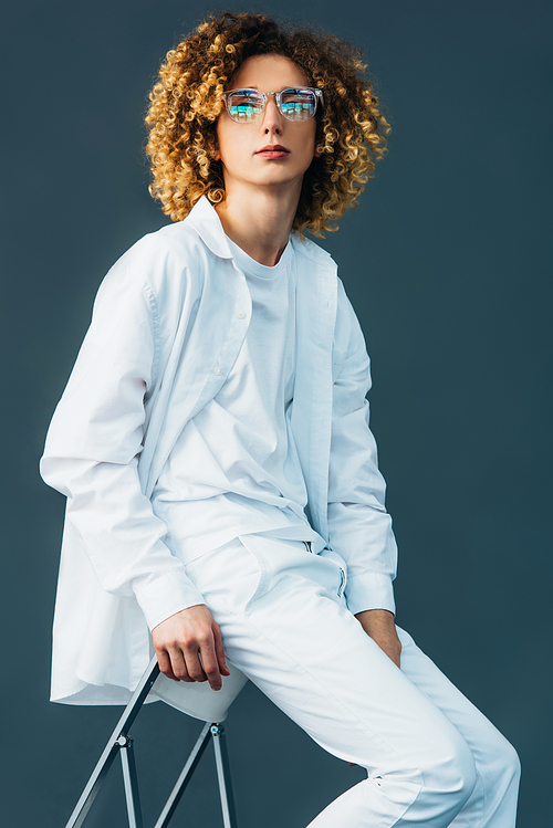 stylish curly teenager in total white outfit and glasses sitting on chair isolated on green