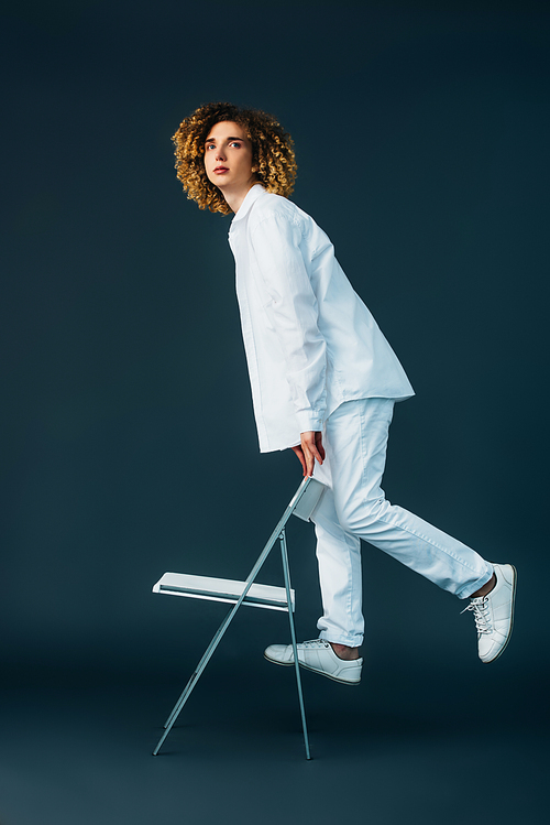 stylish curly teenager in total white outfit standing on chair on green