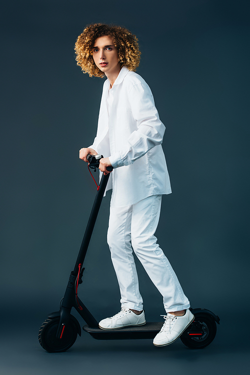 stylish curly teenager in total white outfit riding electric scooter on green