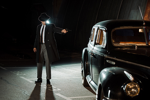 handsome gangster in hat and coat holding gun near retro car