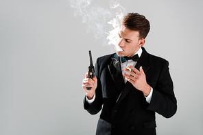 dangerous man in suit with bow tie holding gun while smoking cigar isolated on grey