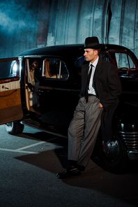 handsome gangster in hat standing with hands in pocket near car