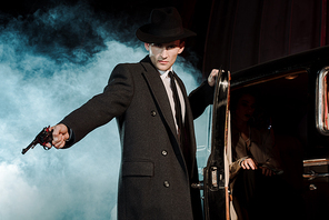 dangerous man in hat holding gun near retro car on black with smoke