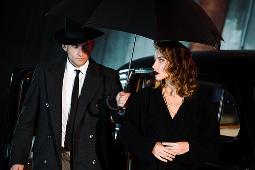 handsome man in hat holding umbrella near attractive girl and vintage car