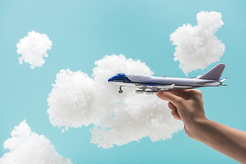 cropped view of woman playing with toy plane among white fluffy clouds made of cotton wool isolated on blue