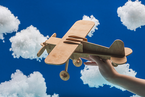 cropped view of woman holding wooden toy plane near white fluffy clouds made of cotton wool isolated on blue