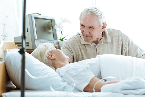 sick senior woman with husband looking at each other in ward in clinic