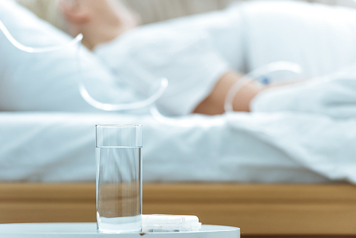 selective focus of sick senior woman lying on bed in clinic with glass of water on foreground