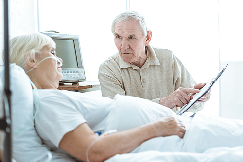 senior man holding clipboard and looking at sick wife in clinic