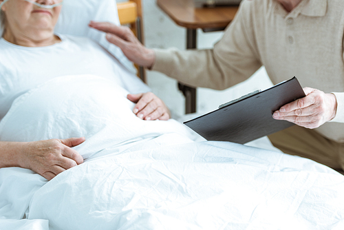 cropped view of senior man holding clipboard with sick wife in clinic