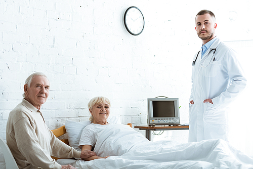doctor in white coat, senior man and smiling ill woman lying on bed in clinic