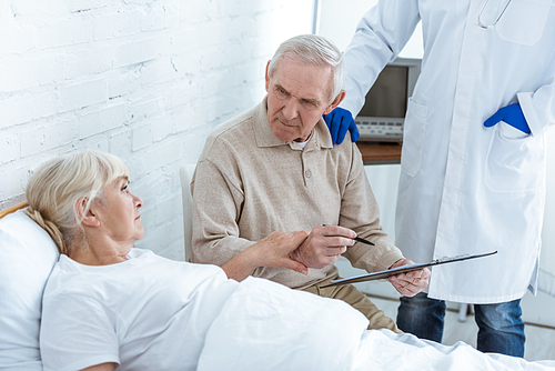 partial view of doctor, senior man signing clipboard and ill patient on bed