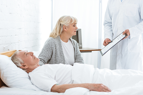 partial view of doctor holding clipboard, sick patient and senior woman in clinic