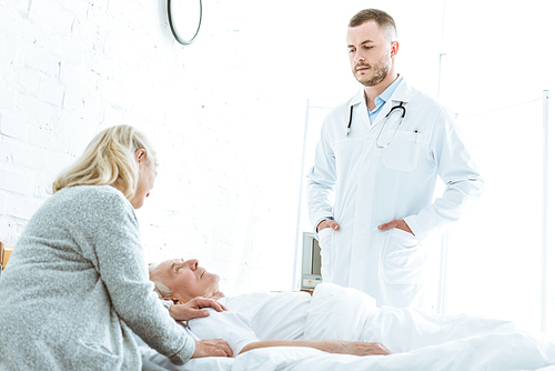 serious doctor standing with hands in pockers near ill patient and senior woman in clinic