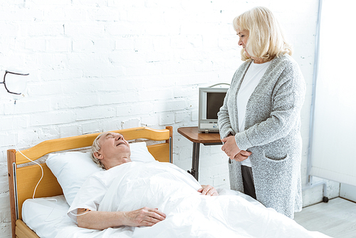 senior woman standing near sick husband in hospital