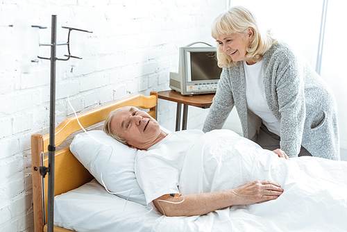 smiling senior woman standing near ill husbend in hospital