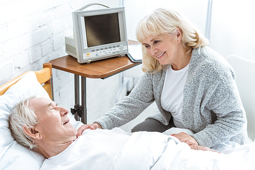 smiling senior woman standing near ill husbend in hospital