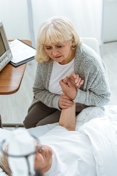 overhead view of worried senior woman sitting near ill husbend and holding his hand in hospital