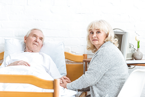 worried senior woman sitting near ill husbend and holding his hand in hospital