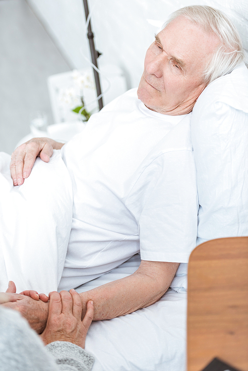 cropped view of senior woman holding hands with sick husband in hospital