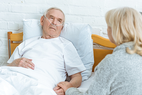 senior woman holding hands with sick husband lying on bed in clinic