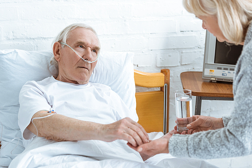 cropped view of senior woman giving glass of water to sick husband in hospital