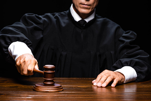 cropped view of judge in judicial robe sitting at table and hitting with gavel isolated on black