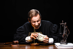 smiling judge in judicial robe sitting at table and holding dollar banknotes isolated on black