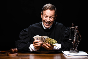 smiling judge in judicial robe sitting at table and holding dollar banknotes isolated on black