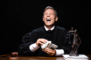 smiling judge in judicial robe sitting at table and holding dollar banknotes isolated on black