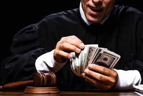 cropped view of shocked judge in judicial robe sitting at table and counting dollar banknotes isolated on black