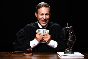 smiling judge in judicial robe sitting at table and holding dollar banknotes isolated on black