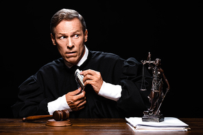 scared judge in judicial robe sitting at table and holding dollar banknotes isolated on black