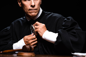 cropped view of scared judge in judicial robe sitting at table and holding dollar banknotes isolated on black